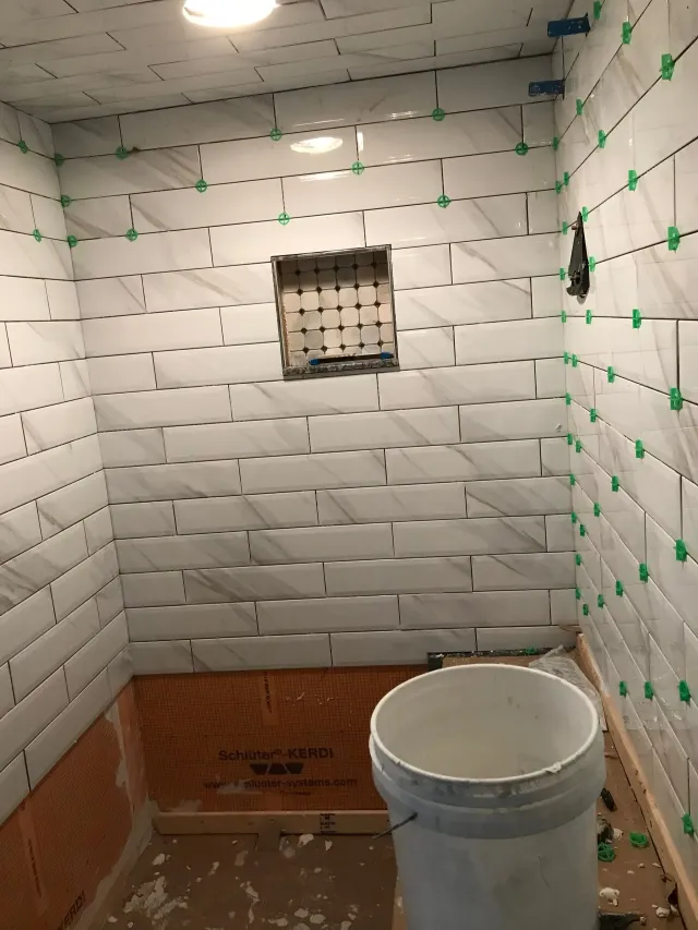 Small tiled bathroom under construction with a bucket on the floor and a vent opening on the back wall