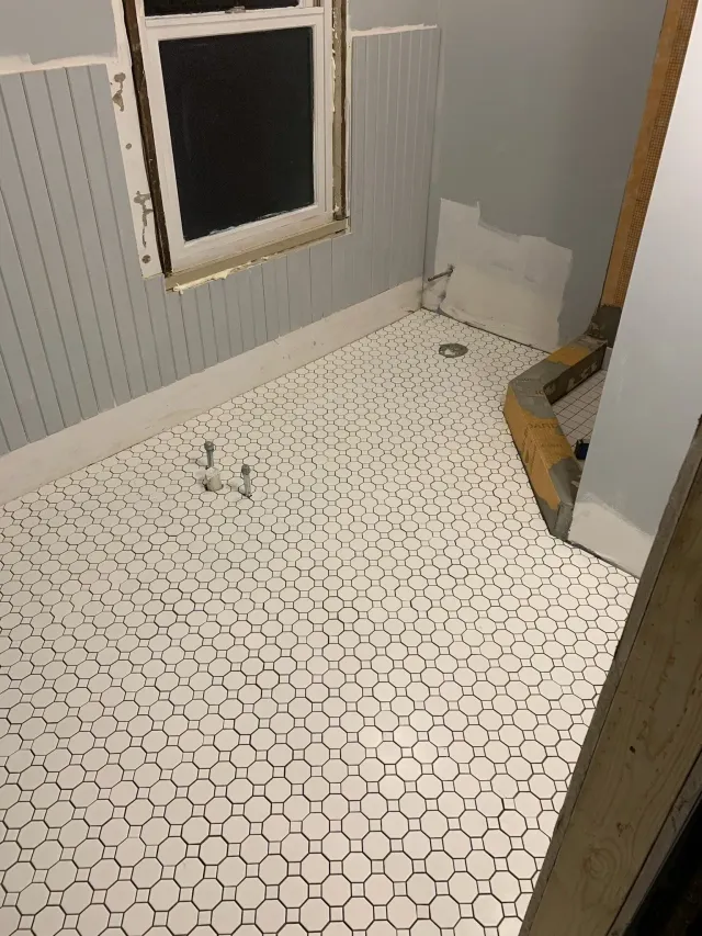 Small bathroom with white patterned floor tiles, gray walls, a window, and a toilet paper holder in the corner.