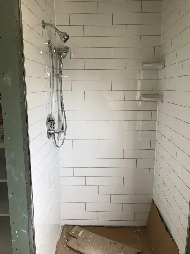 Small white tiled shower stall with exposed plumbing and unfinished floor, no shower door installed