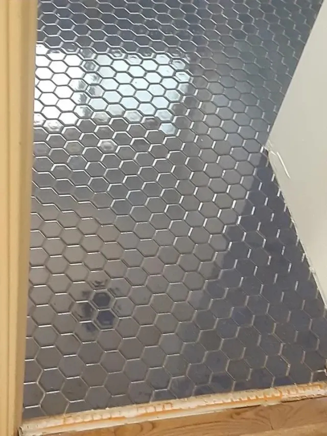 Small bathroom floor and shower wall with dark hexagonal tiles and a bright window reflection