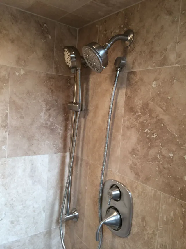 Chrome shower fixture with handheld showerhead and control knob against beige tiled wall