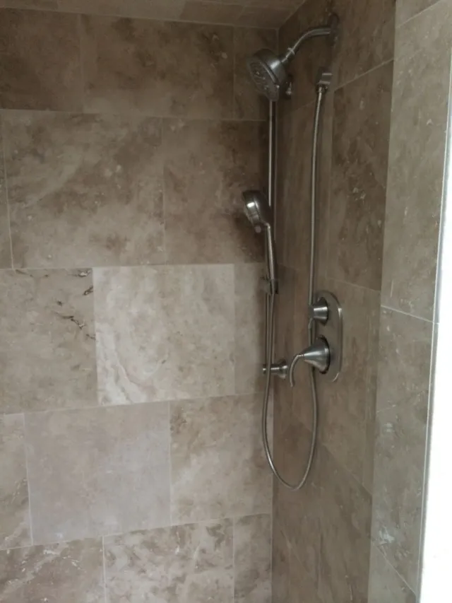 Tiled shower corner with chrome showerhead, hose, and control valve in a beige bathroom