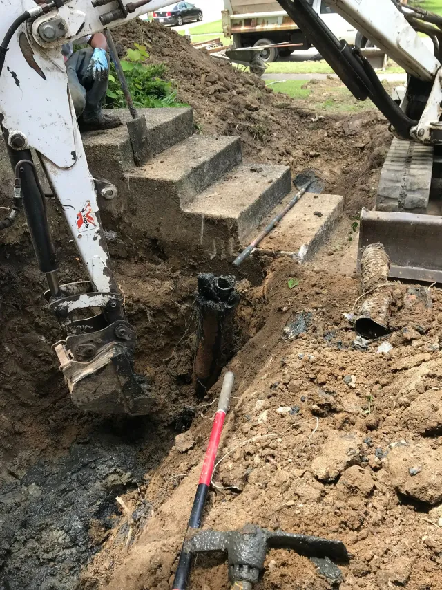 Excavator digging soil beside concrete steps, with muddy ground and exposed pipes nearby.
