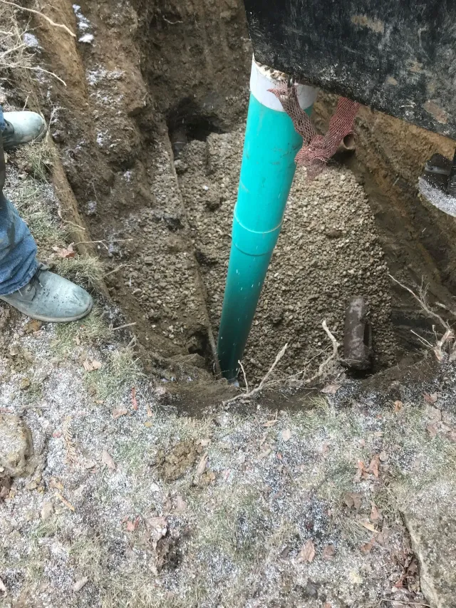 Person installing a green pipe in a dug-up dirt trench beside a booted worker’s feet