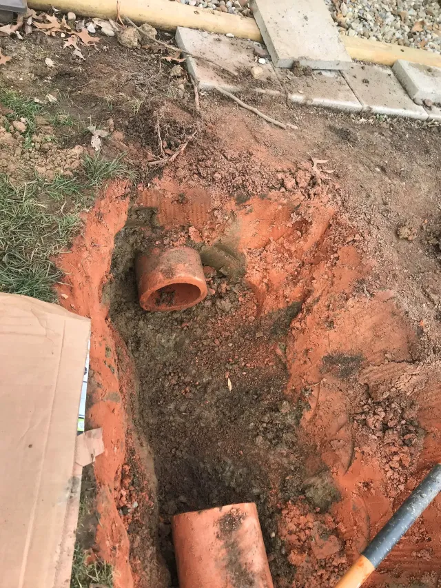 Two exposed orange PVC pipe openings in a dirt trench beside a concrete edge.