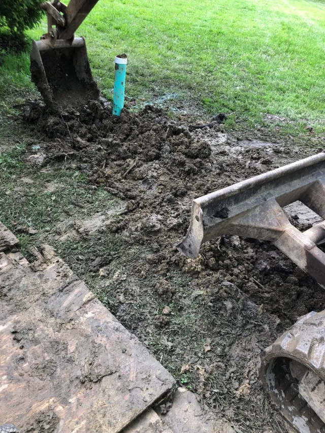 Muddy construction area with a turquoise pipe, dirt pile, and excavator bucket beside grass