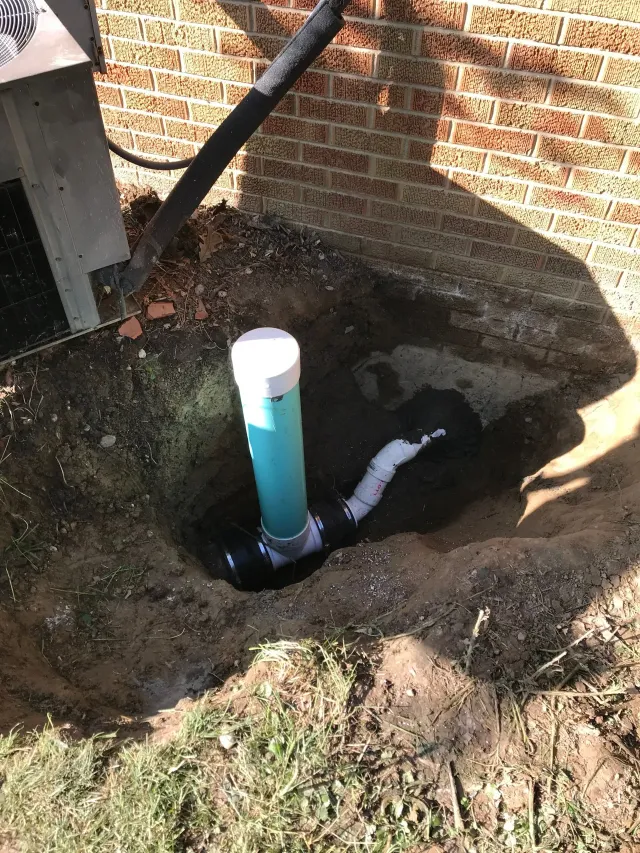 A dug-up yard with a blue PVC pipe standing in a dirt hole beside a brick wall.