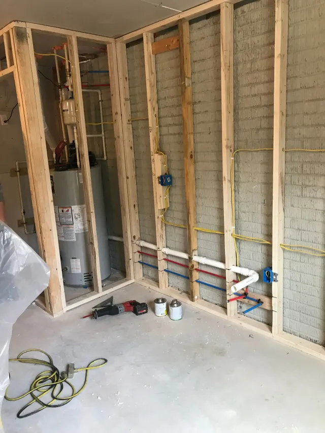 Unfinished interior wall framing with exposed insulation, wiring, and utility connections in a room under construction